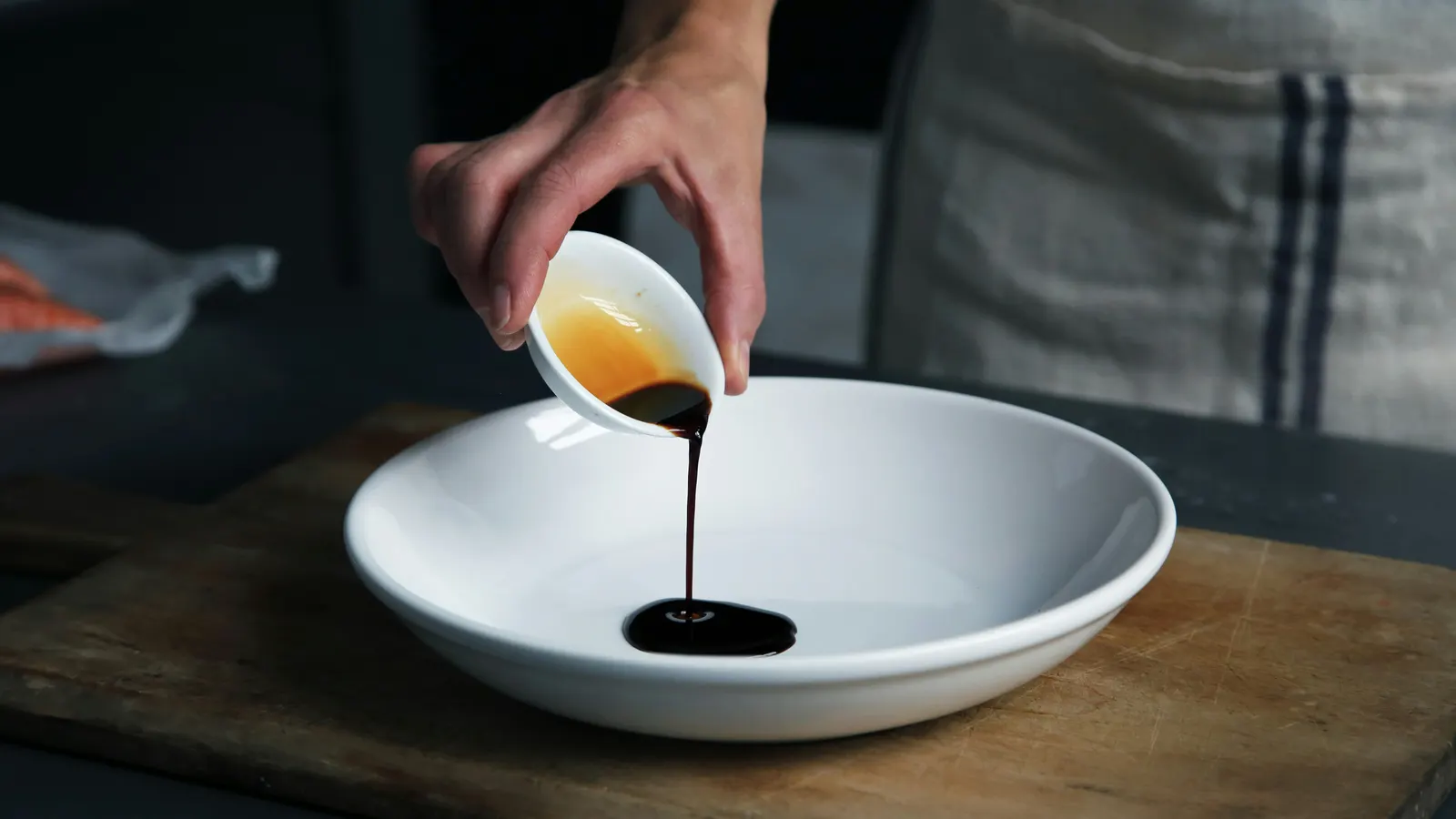 A hand pouring dark balsamic-style vinegar from a small white ceramic bowl into a large white plate, on a wooden cutting board with a striped kitchen towel in the background