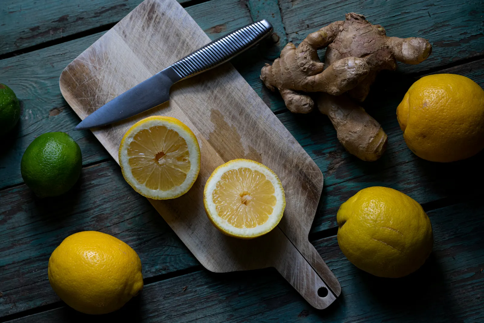 Halved lemons, a whole lemon, a lime, and a piece of fresh ginger arranged on a wooden cutting board with a chef's knife, photographed from above on a dark blue-painted wooden surface