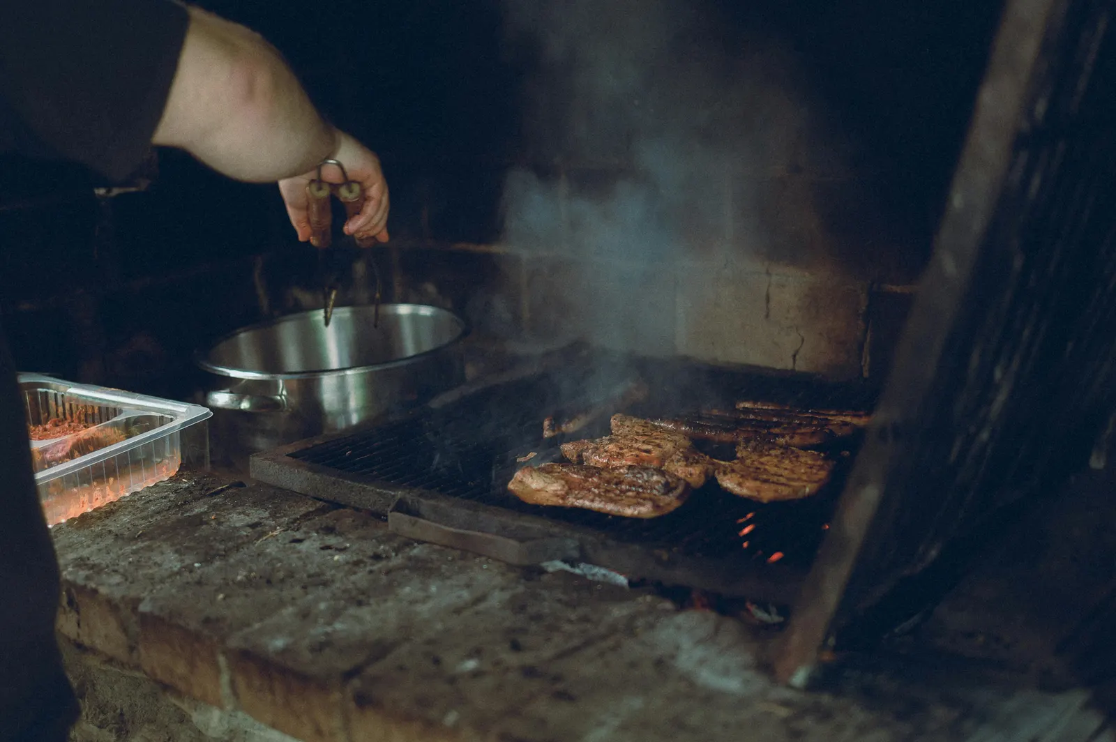 A chef using tongs to turn steaks on a hot charcoal grill, smoke rising in the warm evening light