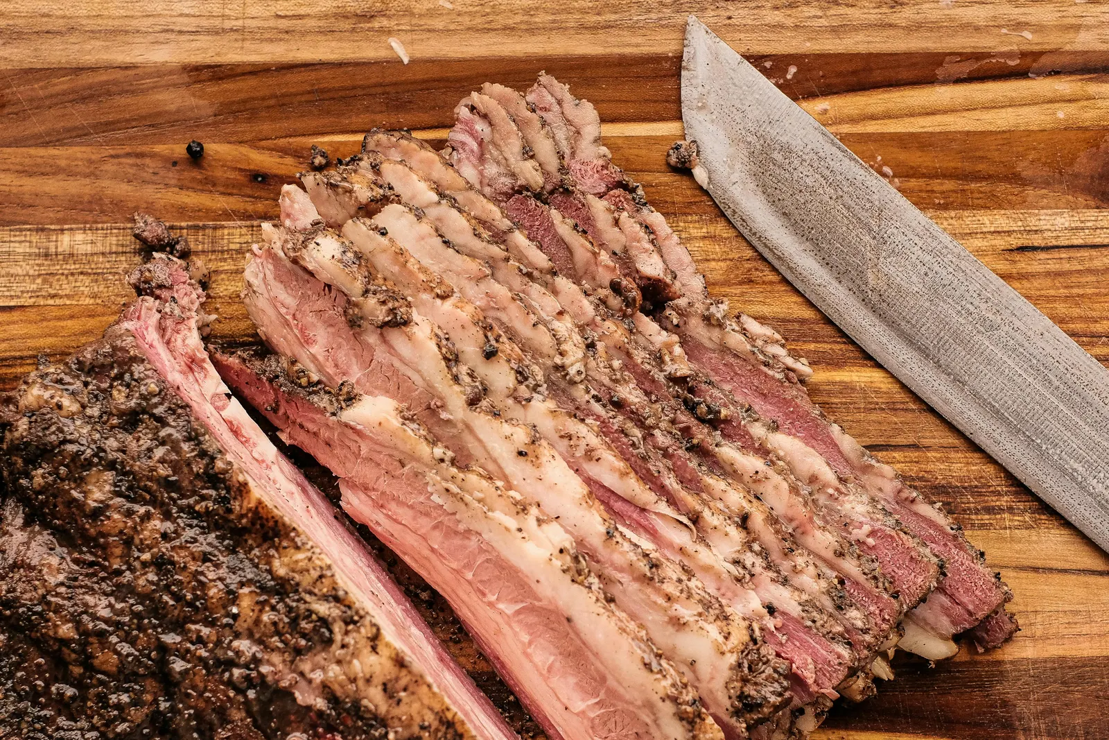 A sliced cooked brisket fanned out on a wooden cutting board, the bark dark and the interior pink, a sharp knife alongside