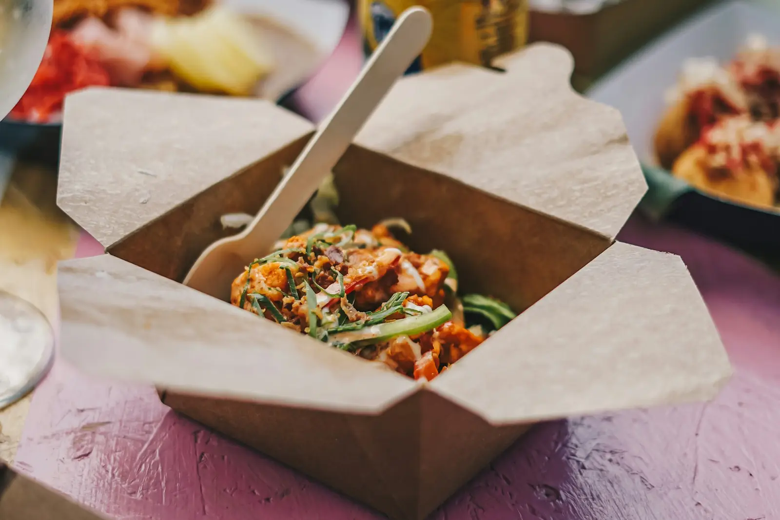 An open kraft takeout container of saucy noodles and vegetables on a table with other dishes around it
