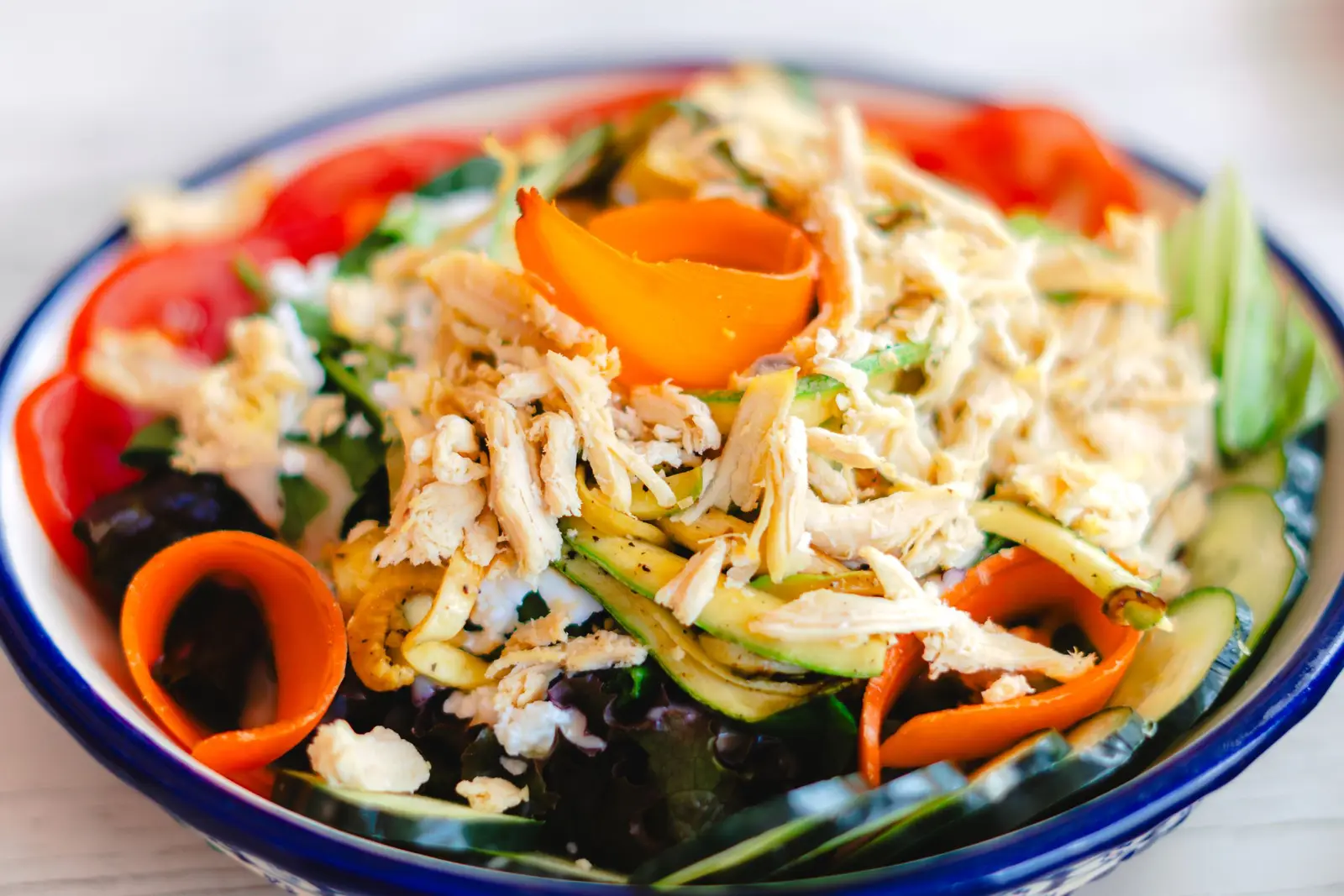 Shredded confit chicken over mixed greens, cucumber, and tomatoes in a blue-rimmed bowl