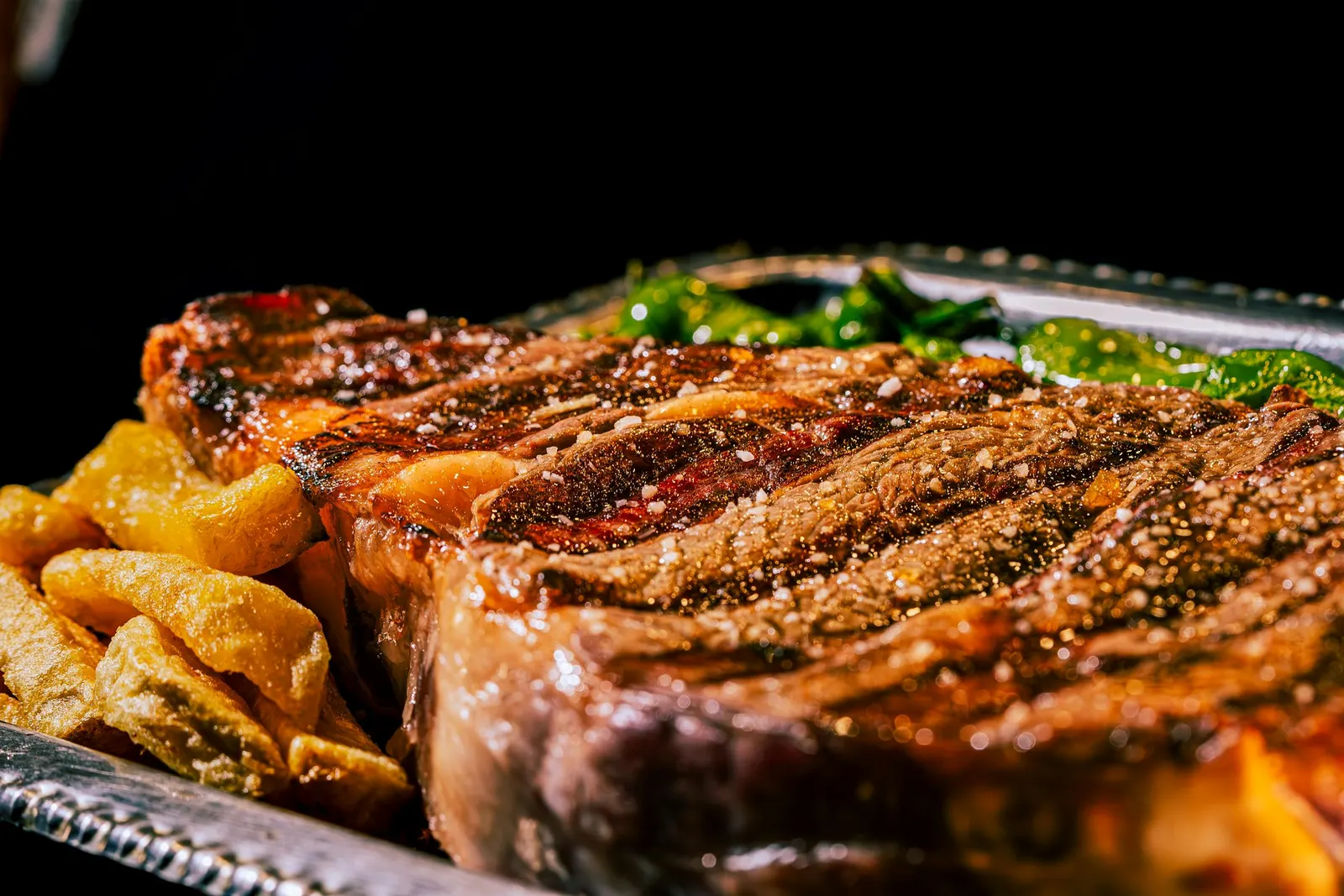 A grilled ribeye steak with a dark seared crust and grill marks, the rendered fat glistening, served with fries and greens