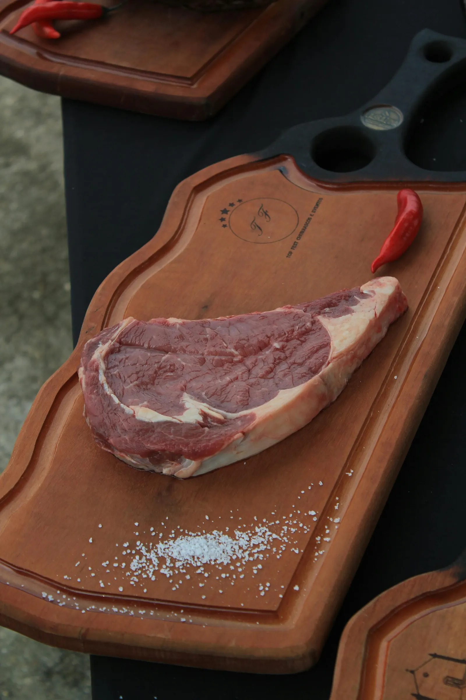A raw ribeye steak on a wooden board showing the central eye and the marbled fat cap, salt and chili alongside