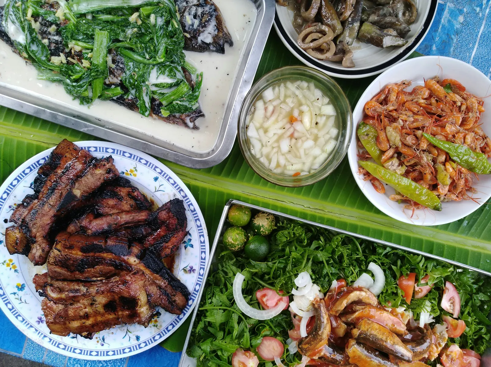 A Filipino home-style spread on a table covered with banana leaves: grilled pork ribs, fried small fish, vinegared vegetables, sautéed water spinach (kangkong), pickled radish, and a salad with calamansi and tomato