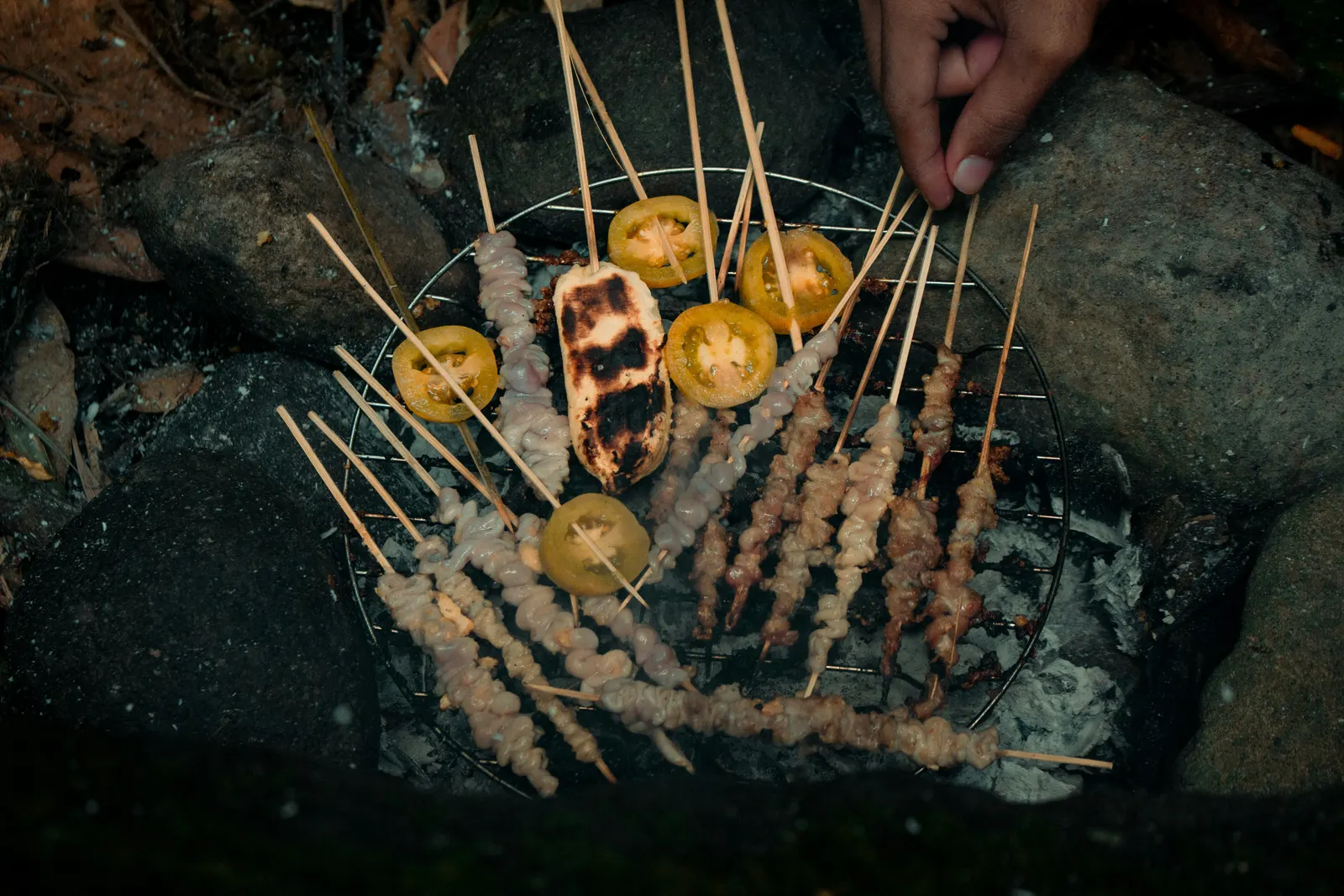 Filipino pork BBQ skewers grilling over charcoal next to slices of yellow tomato, a hand reaching in from the edge of the frame