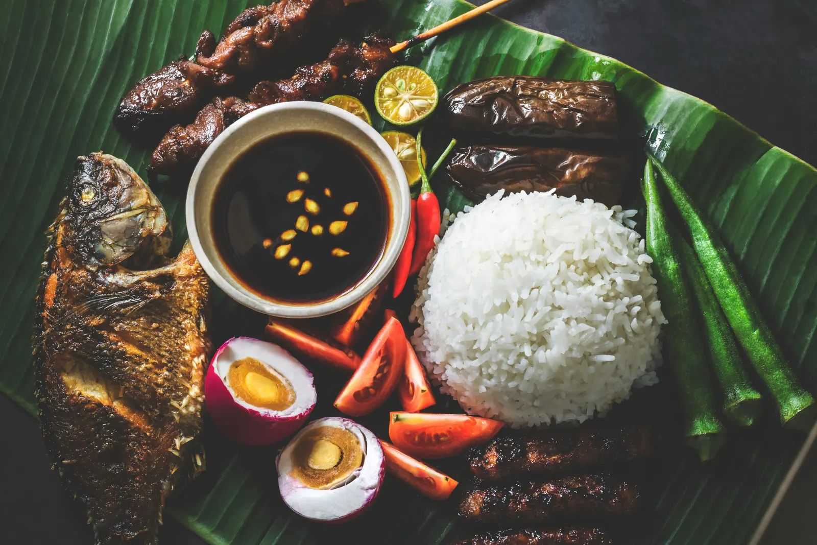An overhead view of a Filipino home-style platter on banana leaves: fried fish, pork BBQ skewers, eggplant, white rice, salted egg, sliced tomato, calamansi, okra, and a small bowl of soy dipping sauce