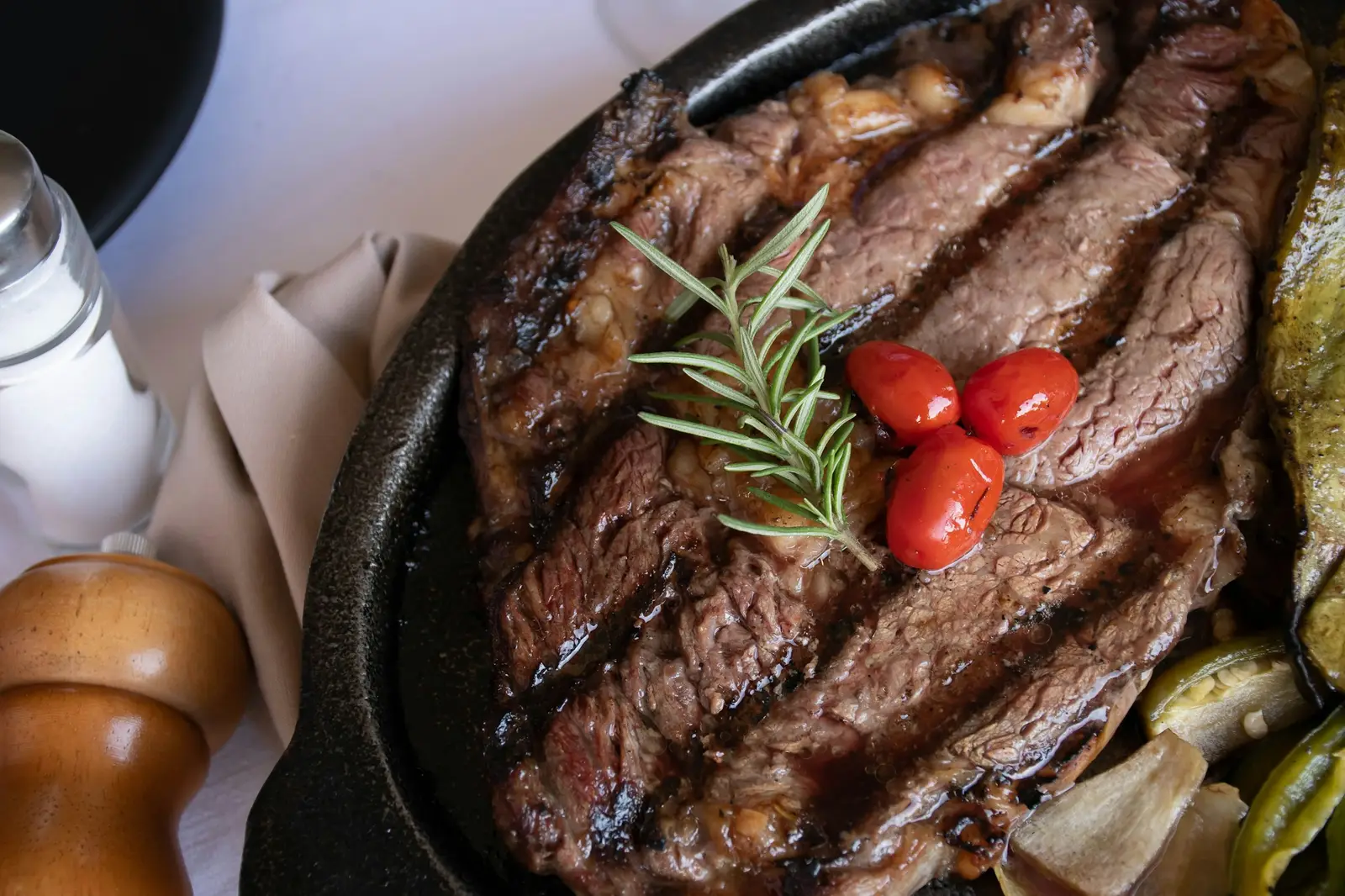 A cooked steak on a dark pan plated with tomato and green vegetables