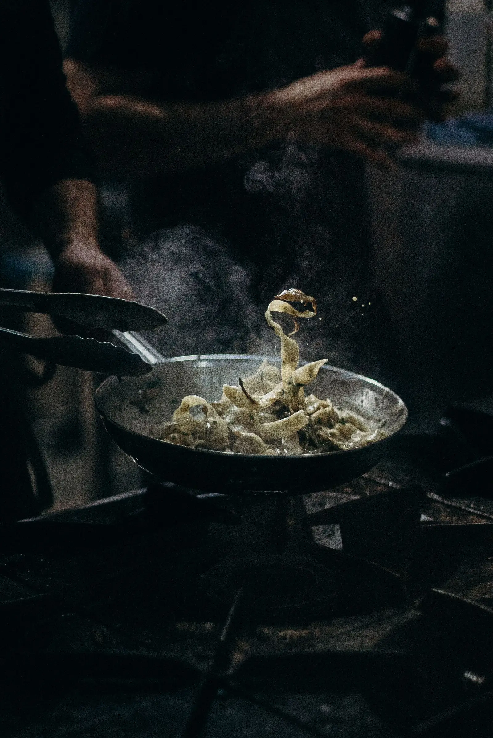 A chef tossing tagliatelle in a steel pan over a flame in a dark restaurant kitchen, steam rising