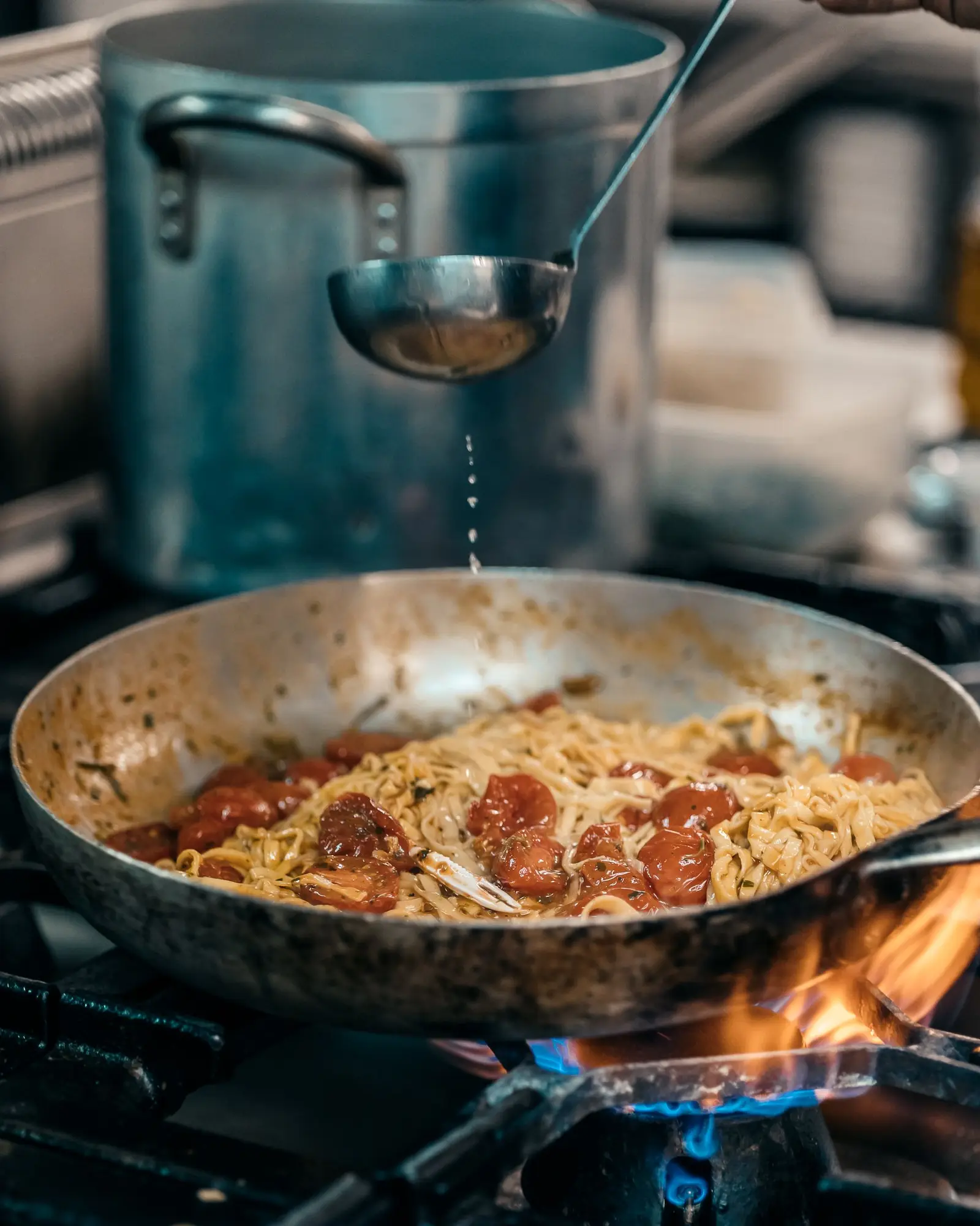 A cook ladling starchy pasta water into a pan of tagliatelle with cherry tomatoes over an open flame