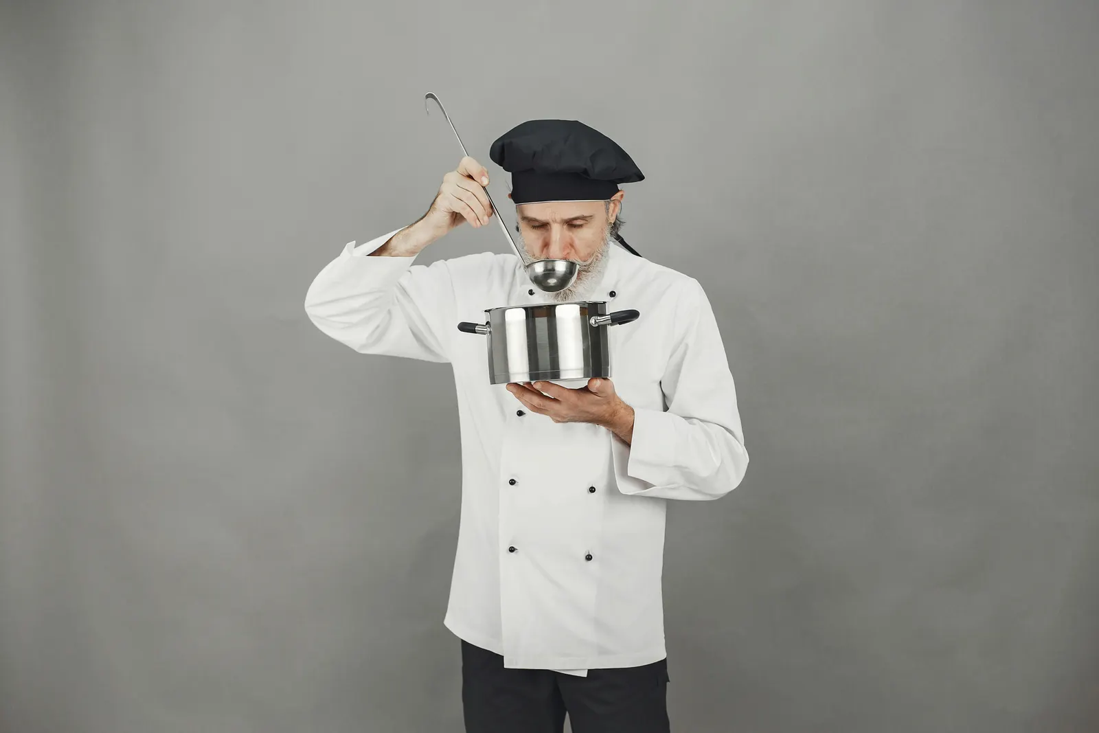 A chef tasting from a wooden spoon held above a pot in a working kitchen