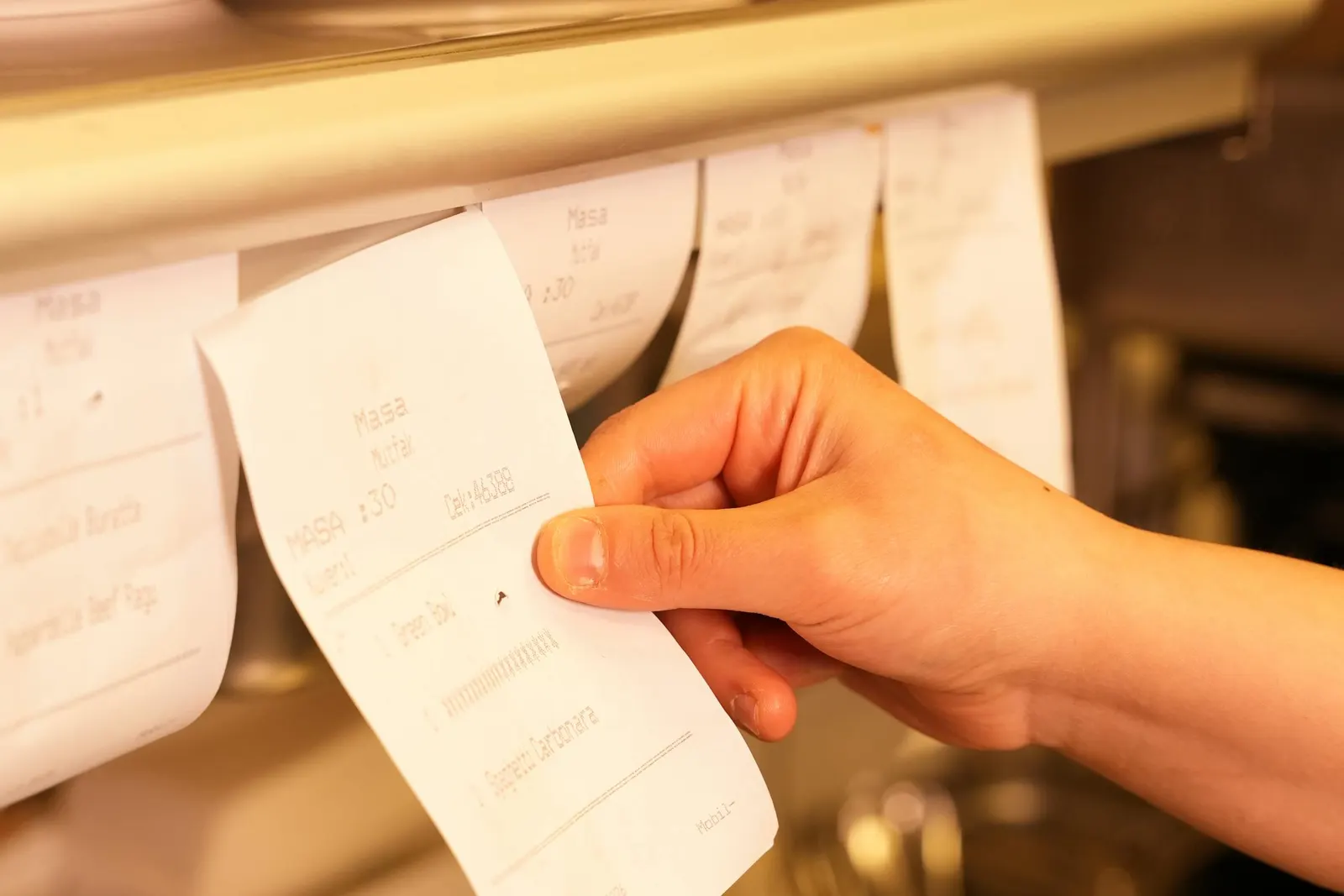 A hand pulling a printed order ticket off the rail at a restaurant kitchen pass, more tickets queued beside it