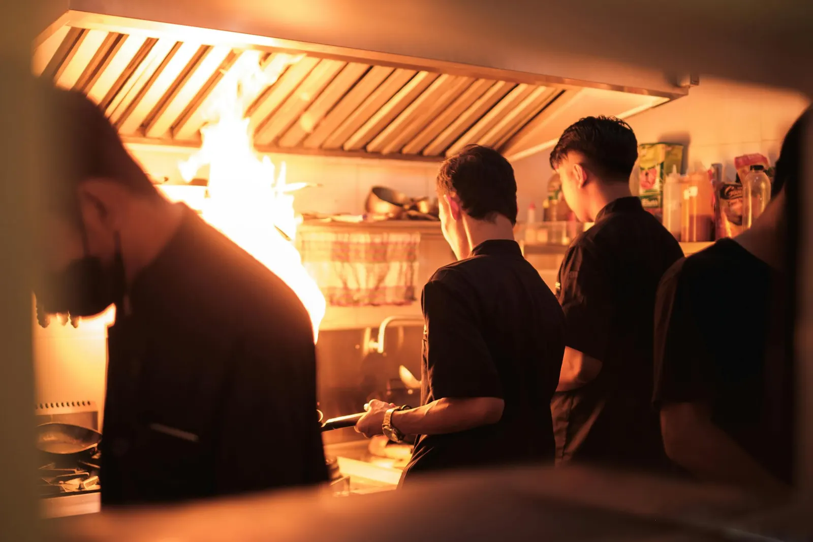 Cooks working a busy restaurant line under a hood vent with a tall flame rising from a sauté pan