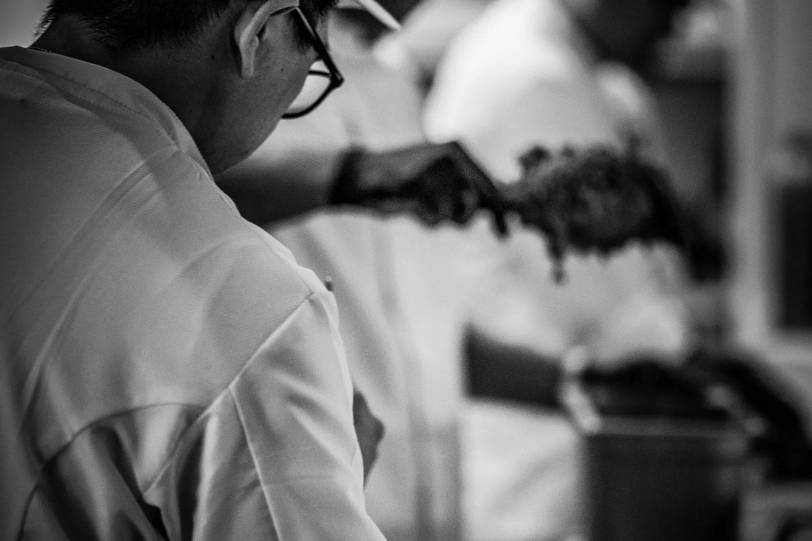 A black and white shot of one chef in focus while another plates a dish with fresh herbs in the background