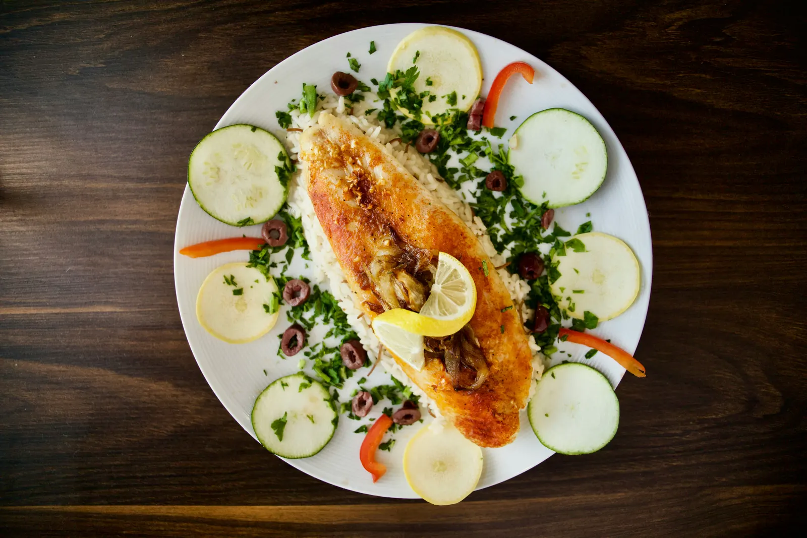 A whole roasted fish on a bed of rice, garnished with parsley, sliced cucumber, olives, lemon wedges, and red pepper, served on a white plate on a dark wooden table
