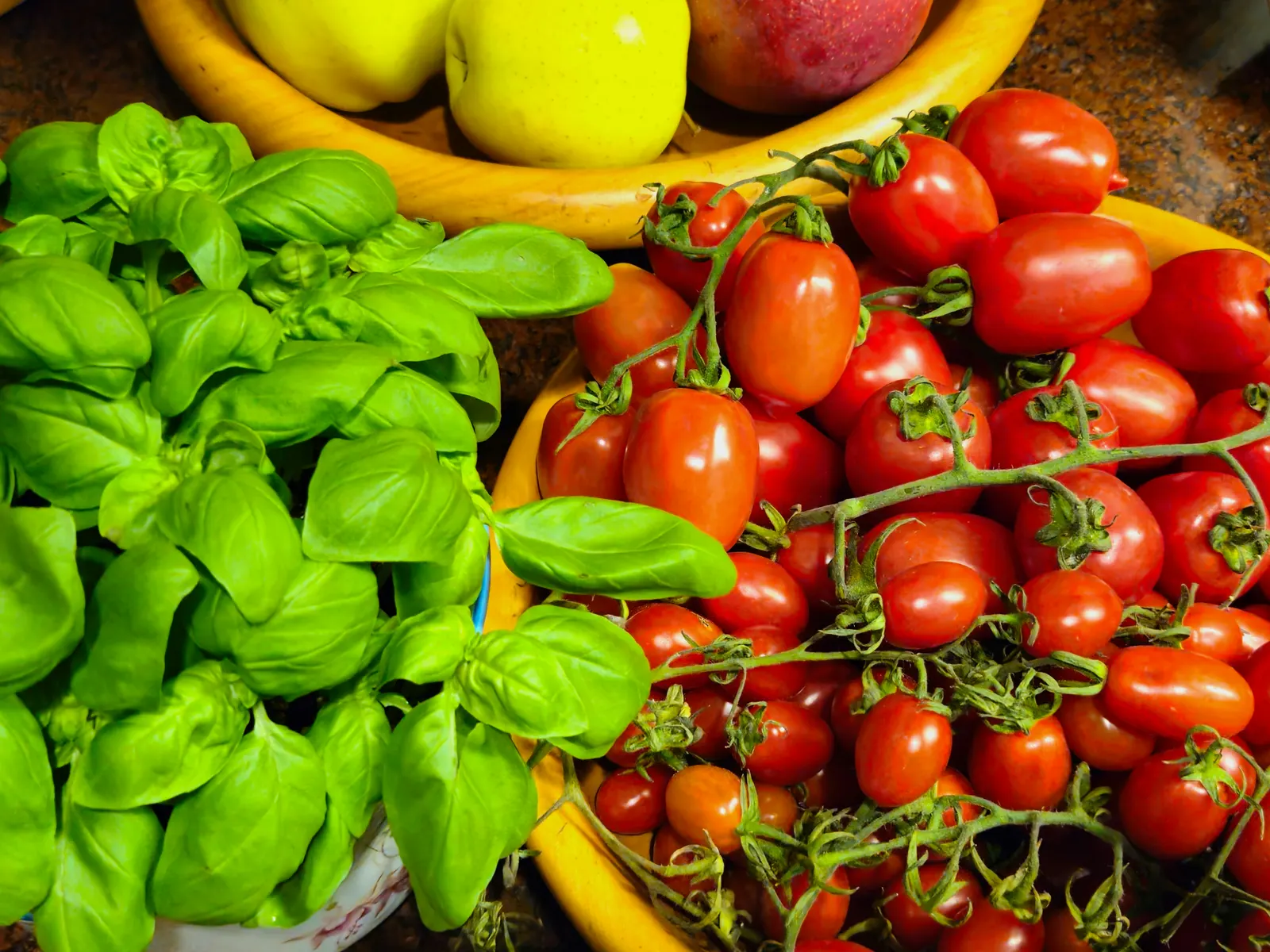 Fresh basil leaves and ripe cherry tomatoes on the vine arranged in two yellow bowls, with apples in the background, a vibrant kitchen still life