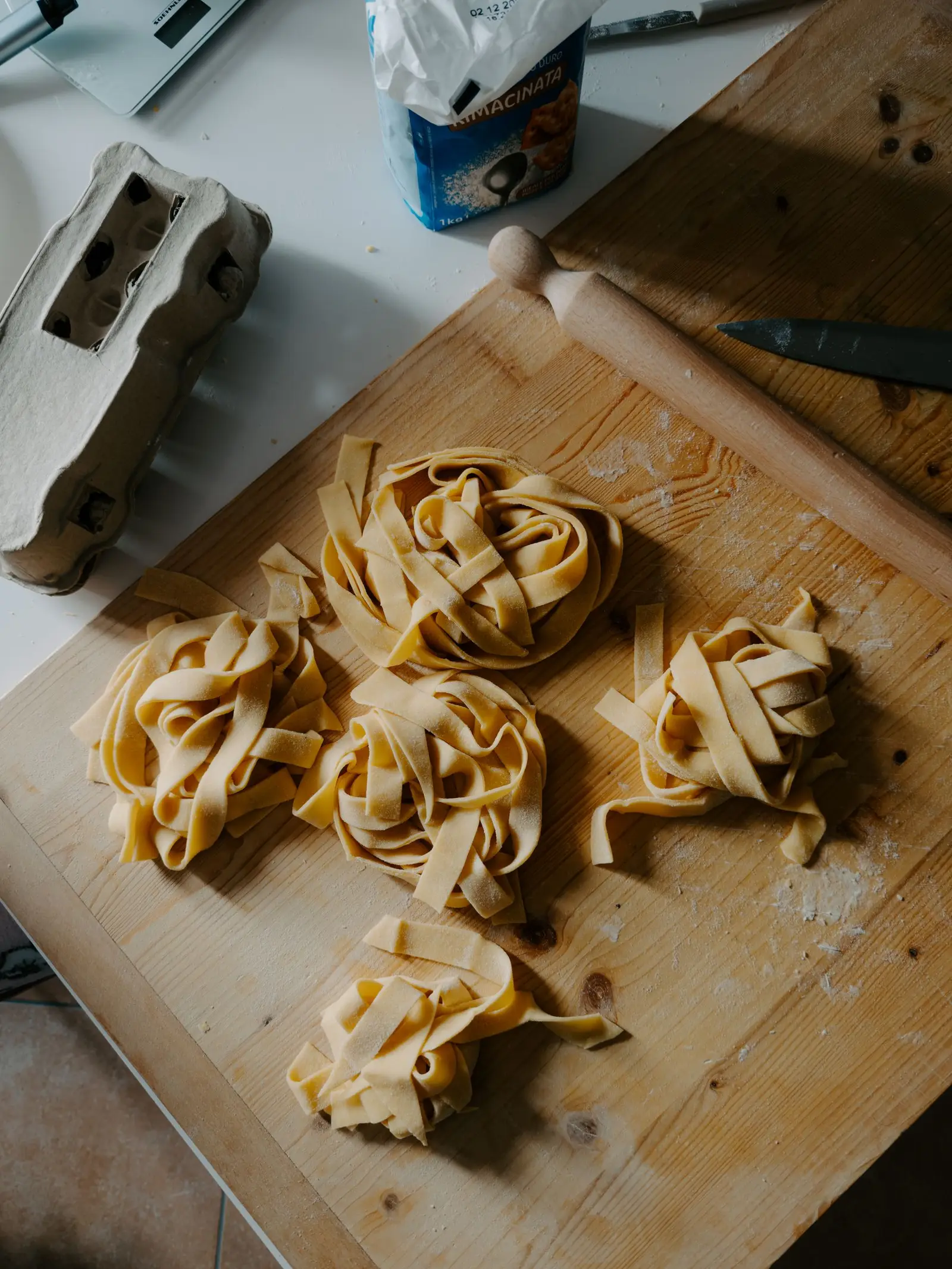 Five nests of fresh egg tagliatelle pasta on a wooden board, with a rolling pin, eggs, and semolina flour