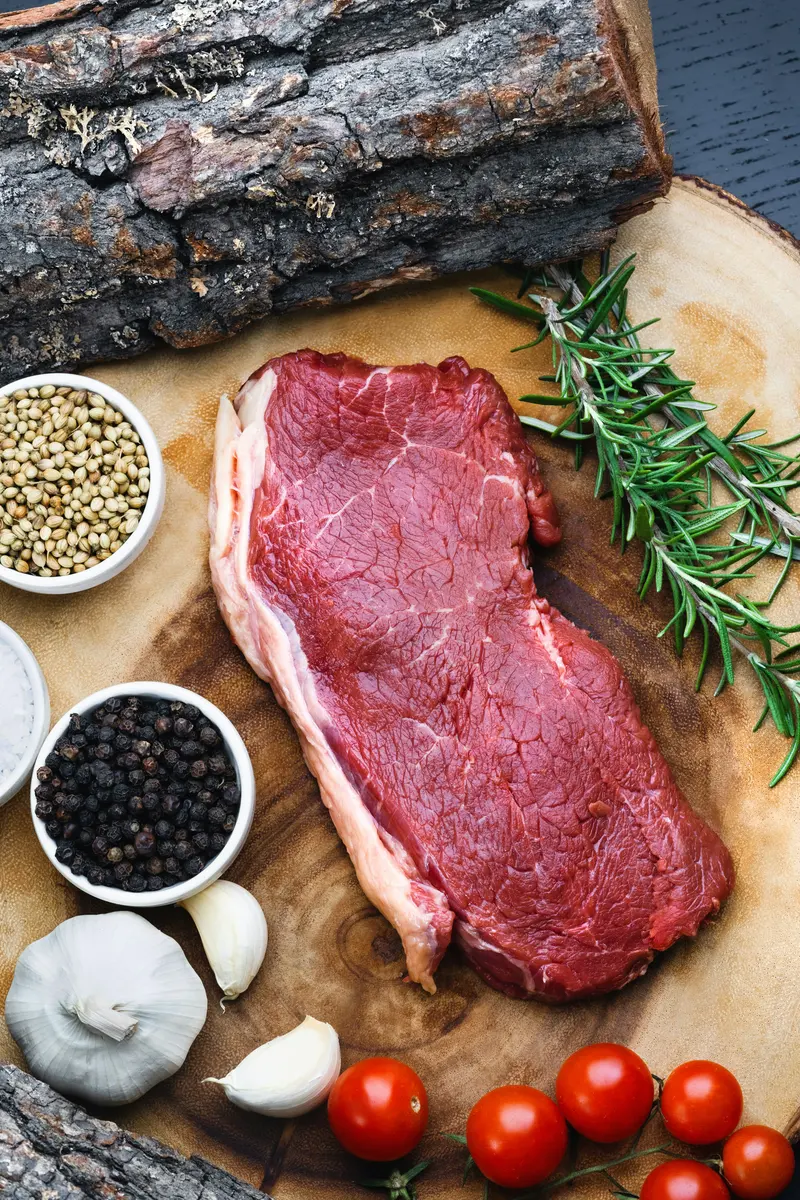 A whole piece of raw beef on a wooden board with peppercorns, garlic, rosemary, and tomatoes, ready to be cut into cubes