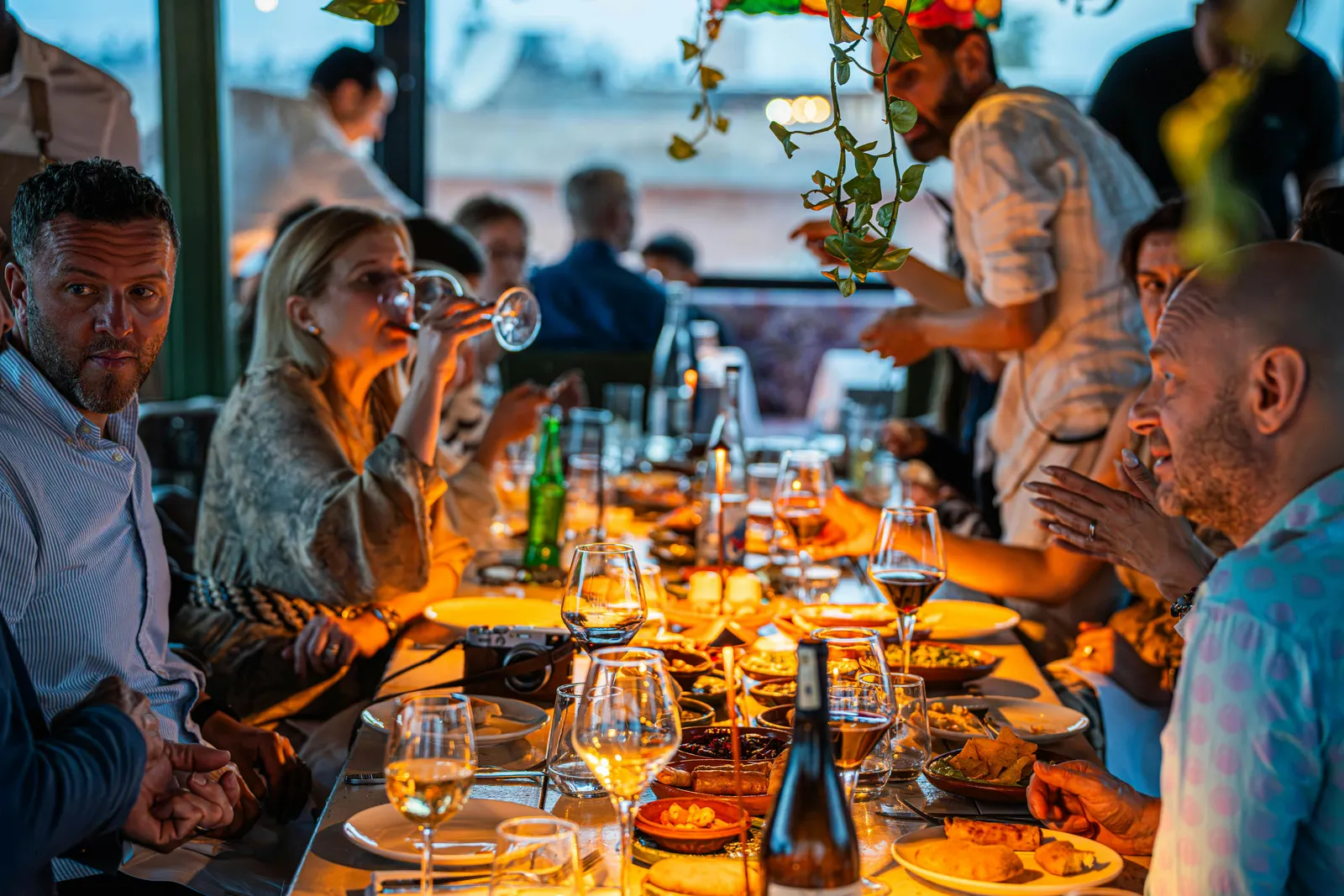 A candlelit restaurant table with friends sharing food and wine in warm low light, the intimate atmosphere of a real night out
