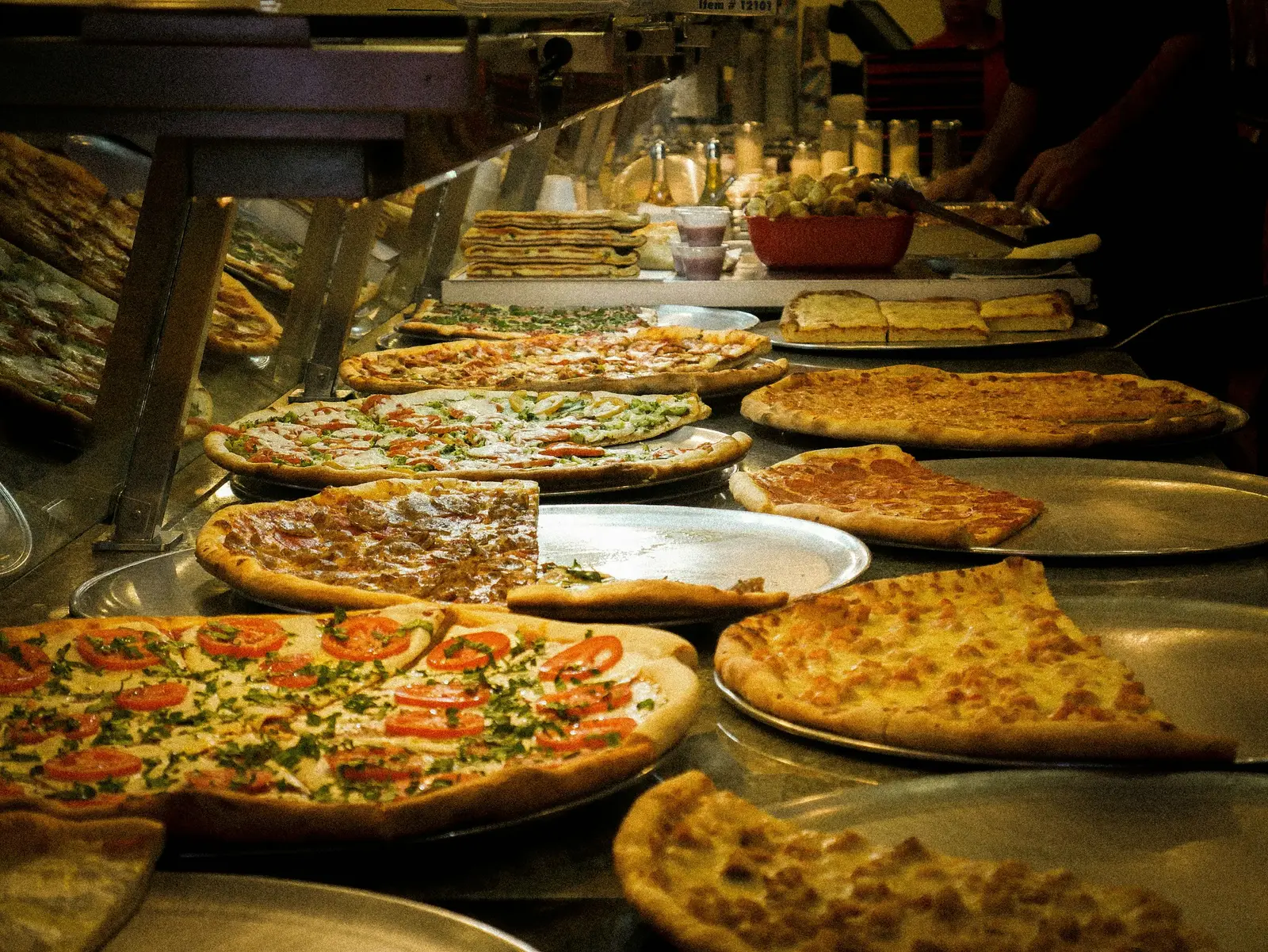 Whole pizzas resting on trays under the warming lights of a New York slice shop counter
