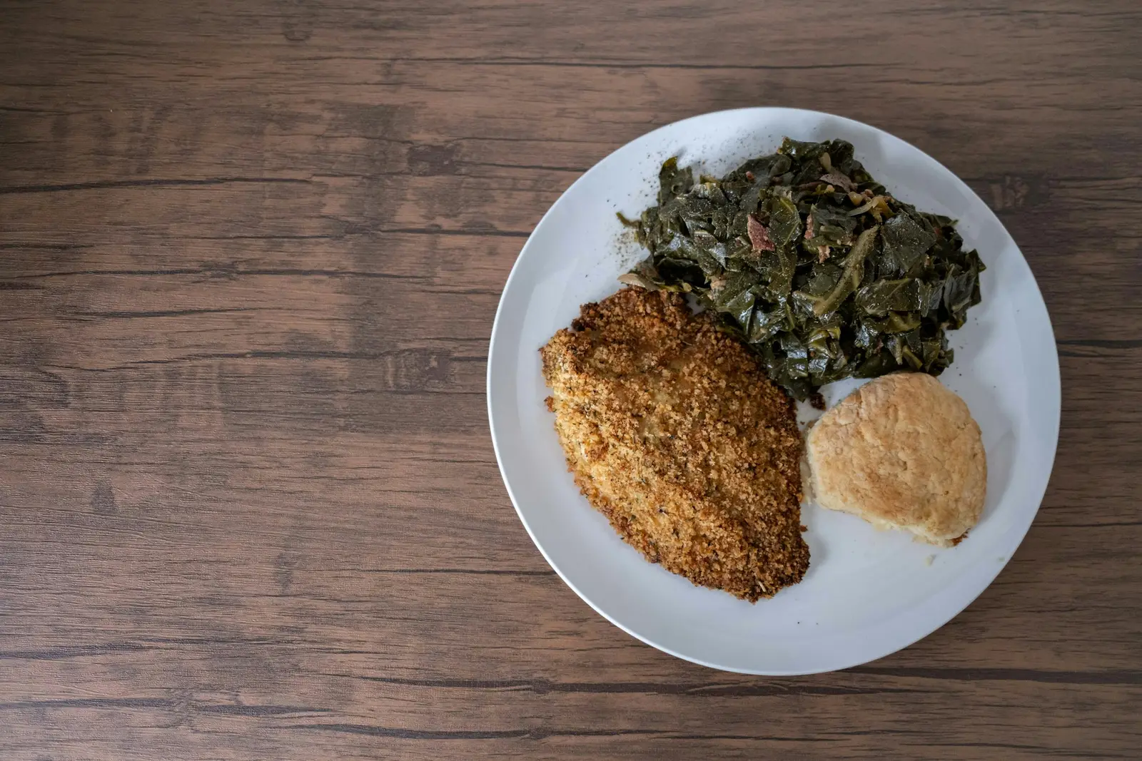 A plate of collard greens, cornbread and fried fish on a dark wooden table