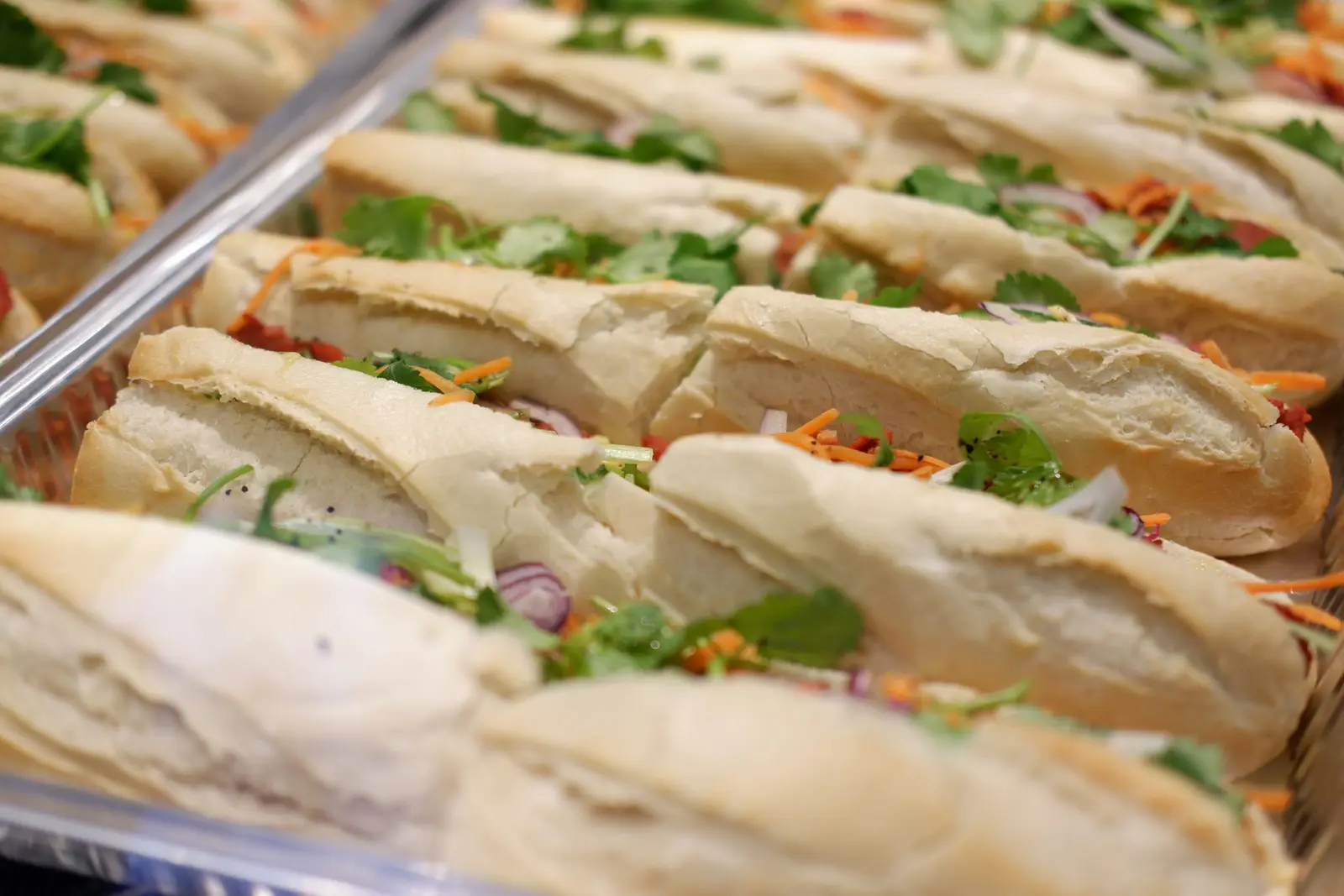 A tray of banh mi baguettes lined up in rows, filled with pickled vegetables, cilantro and chili