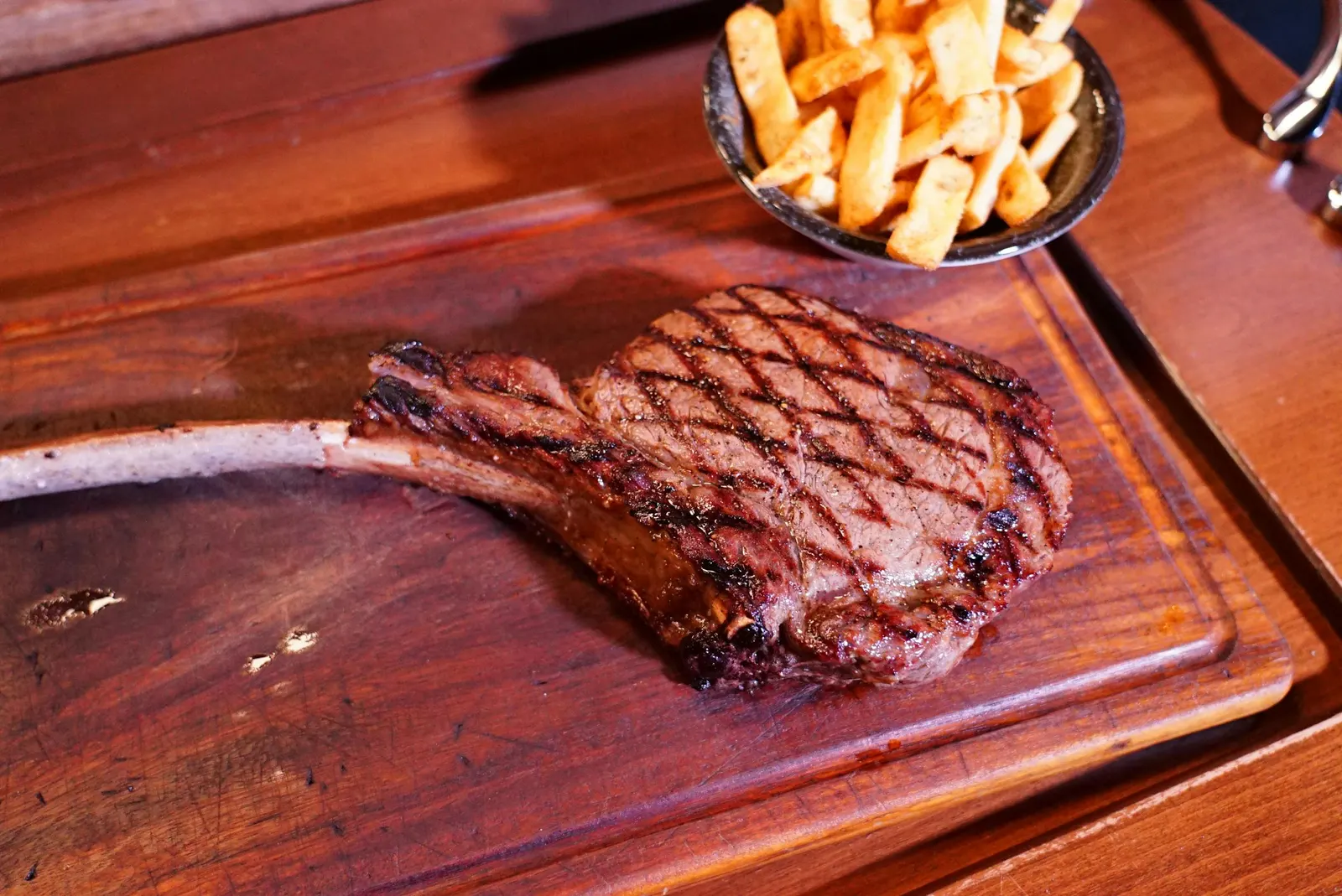 A grilled tomahawk steak with grill marks resting on a wooden board next to a cup of fries, the long bone extending off the board