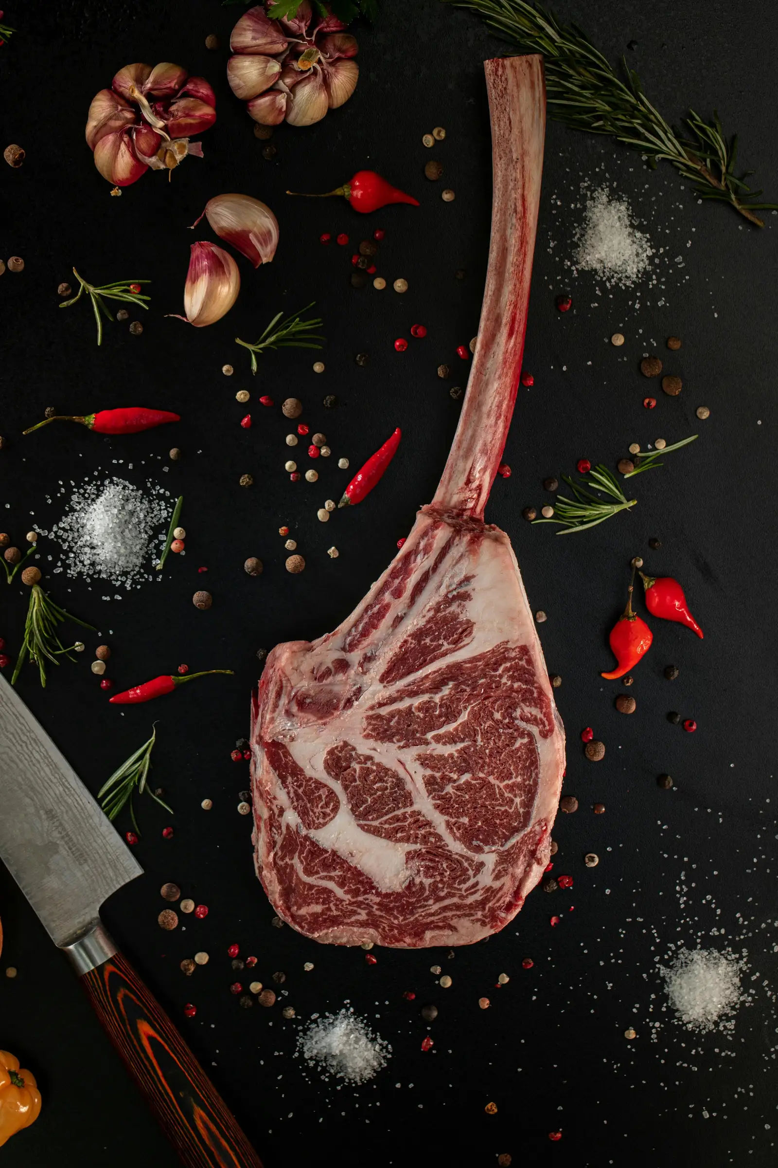 A raw tomahawk steak shot from above on a dark surface, showing heavy marbling in the ribeye and the long frenched bone, surrounded by herbs, garlic, and chili