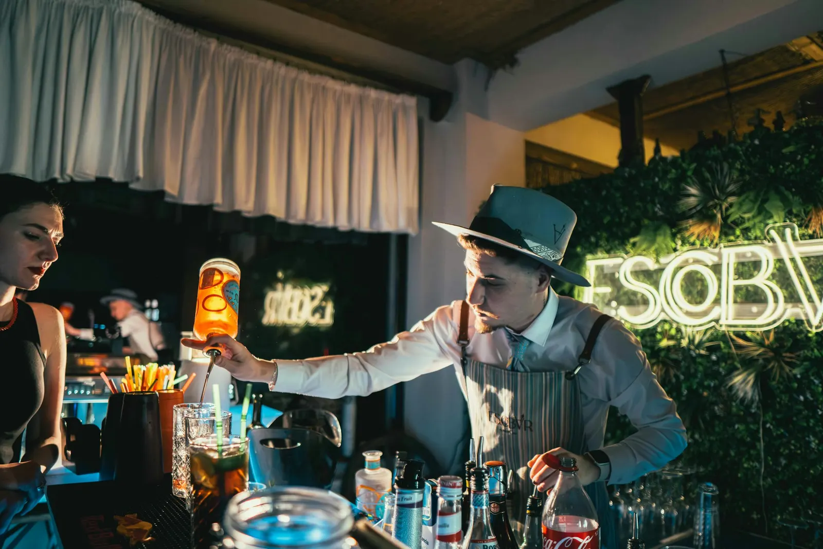 A bartender mid-pour preparing a drink at a dimly lit cocktail bar with neon signage in the background