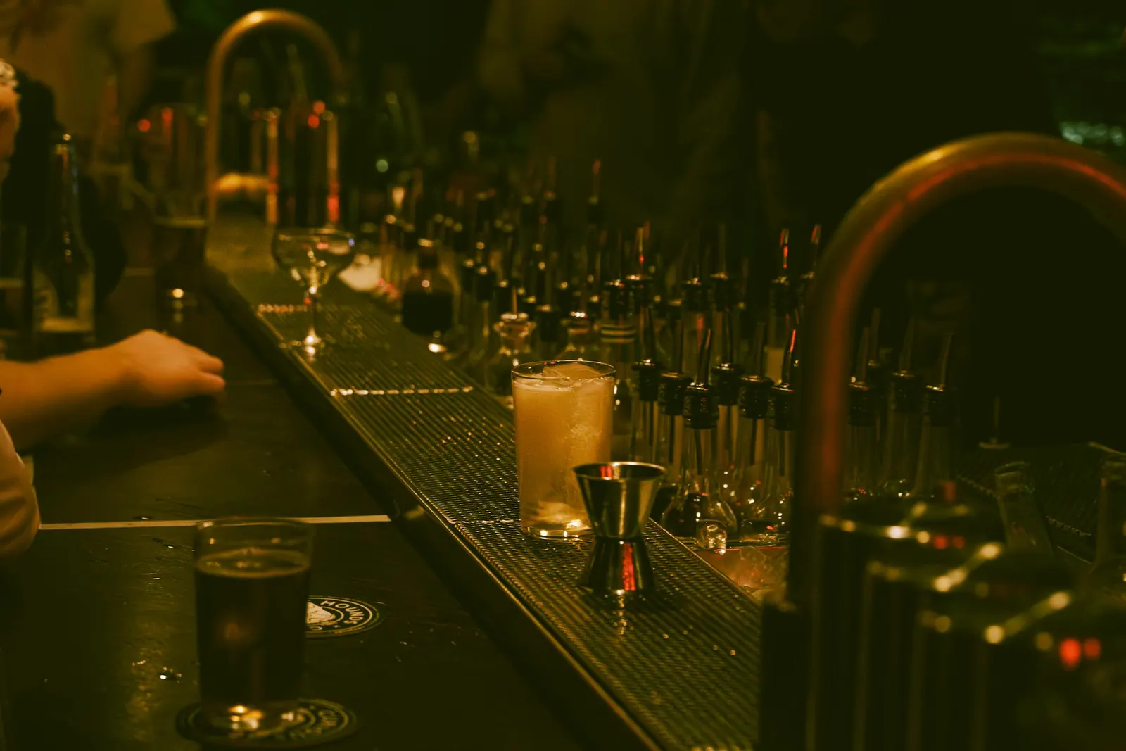 A dimly lit cocktail bar at night with rows of bottles, a glass mid-build, and a hand resting on the counter